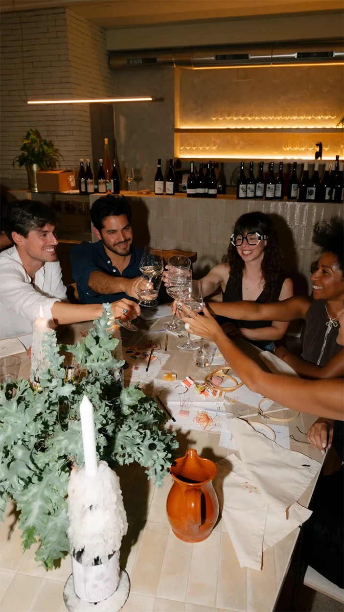 Primer plano de un brindis con copas de vino sobre una mesa de taller del Club Textil de Jugo, rodeada de hilos y materiales de bordado