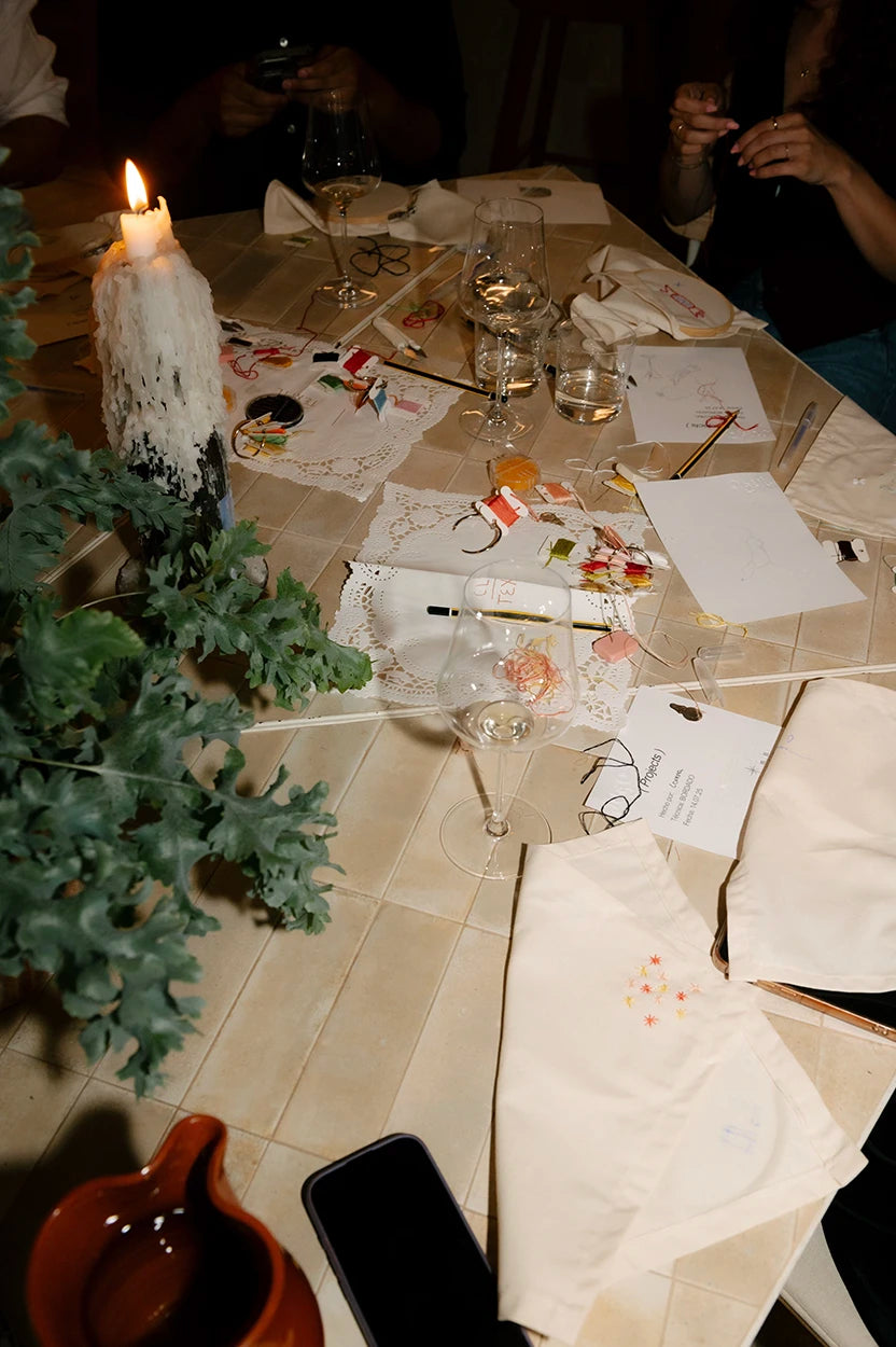 Mesa del Club Textil de Jugo con materiales de bordado, hilos de colores, servilletas hechas a mano, velas y copas de vino