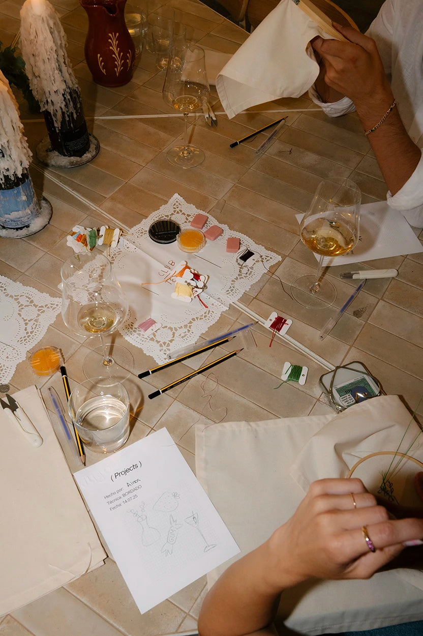 Materiales de bordado sobre la mesa del Club Textil de Jugo: hilos de colores, bastidores de madera, tijeras y copas de vino para una sesión creativa.