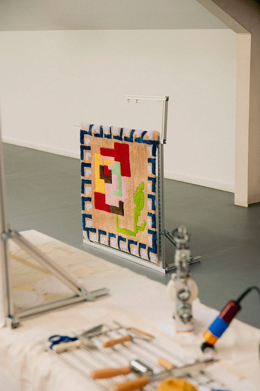 Alfombra de diseño geométrico terminada y expuesta sobre un bastidor metálico en el estudio de Jugo, con herramientas de tufting organizadas en primer plano