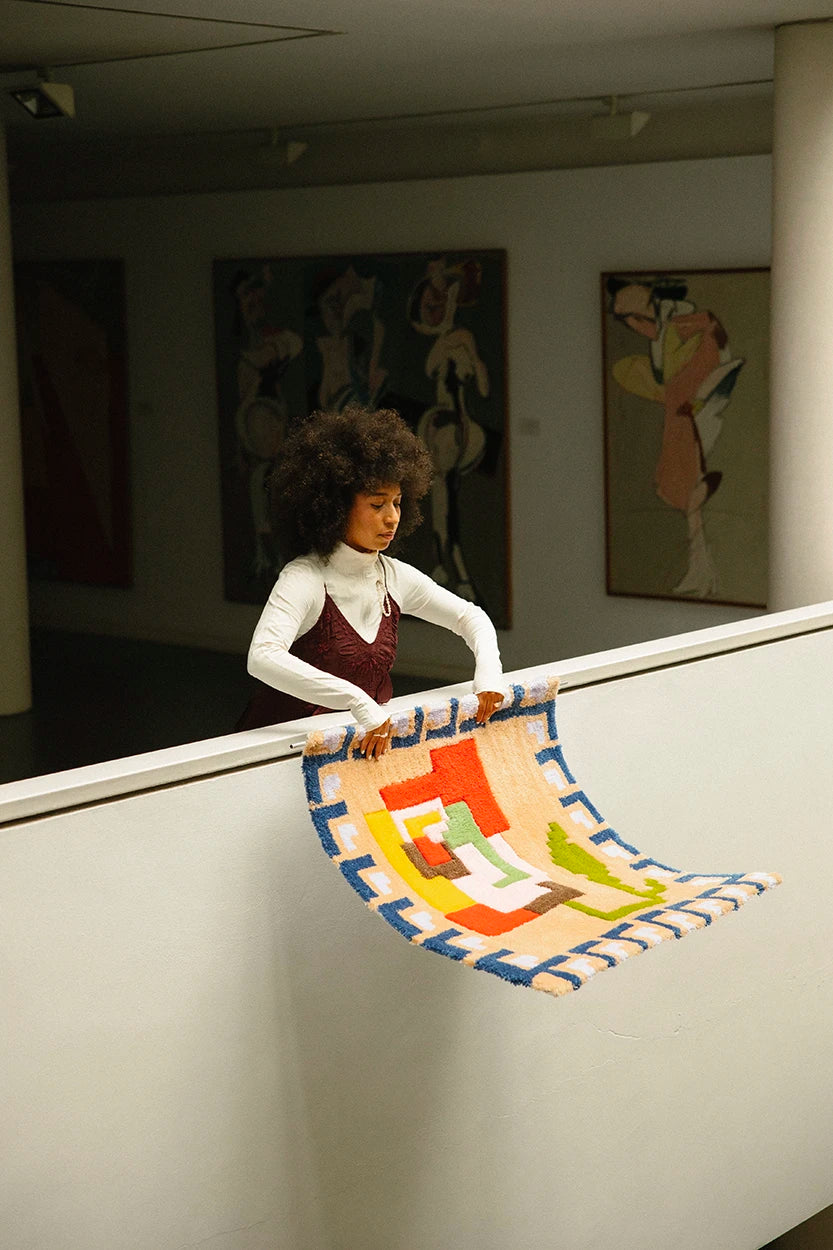 La creadora de Jugo muestra una alfombra artesanal de tufting con diseño geométrico y colorista sobre una barandilla, en un espacio de estudio con arte de fondo.